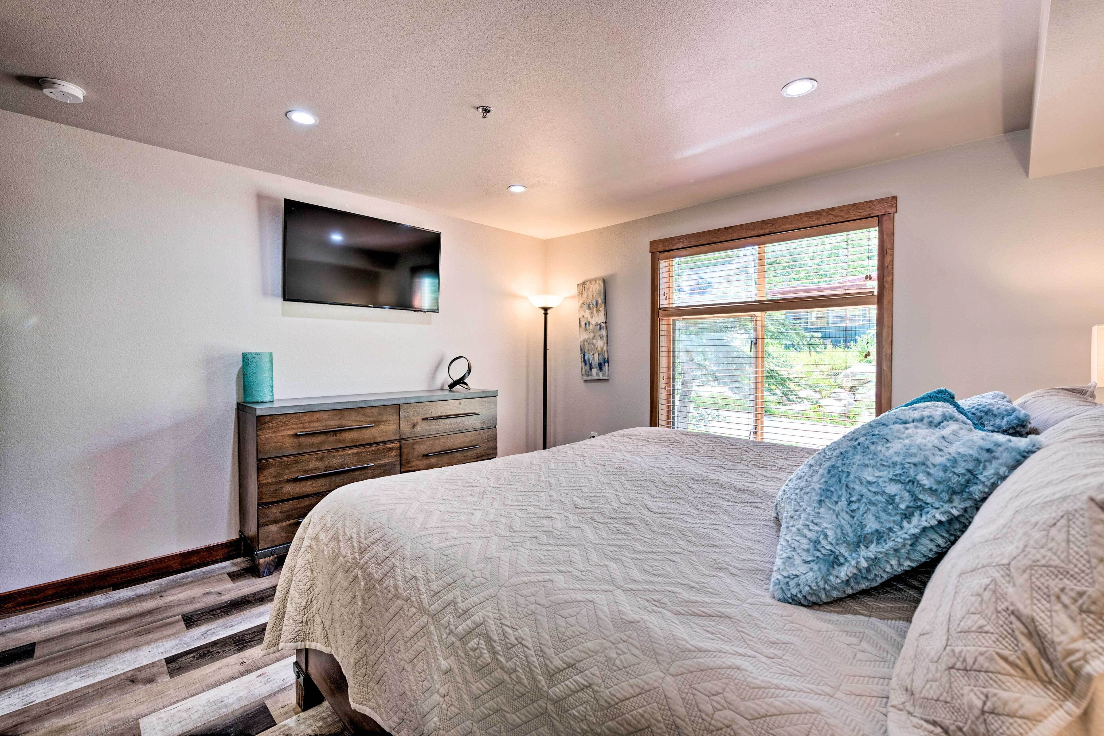 A bedroom with a bed, a dresser, a TV mounted on the wall, and a window with blinds.