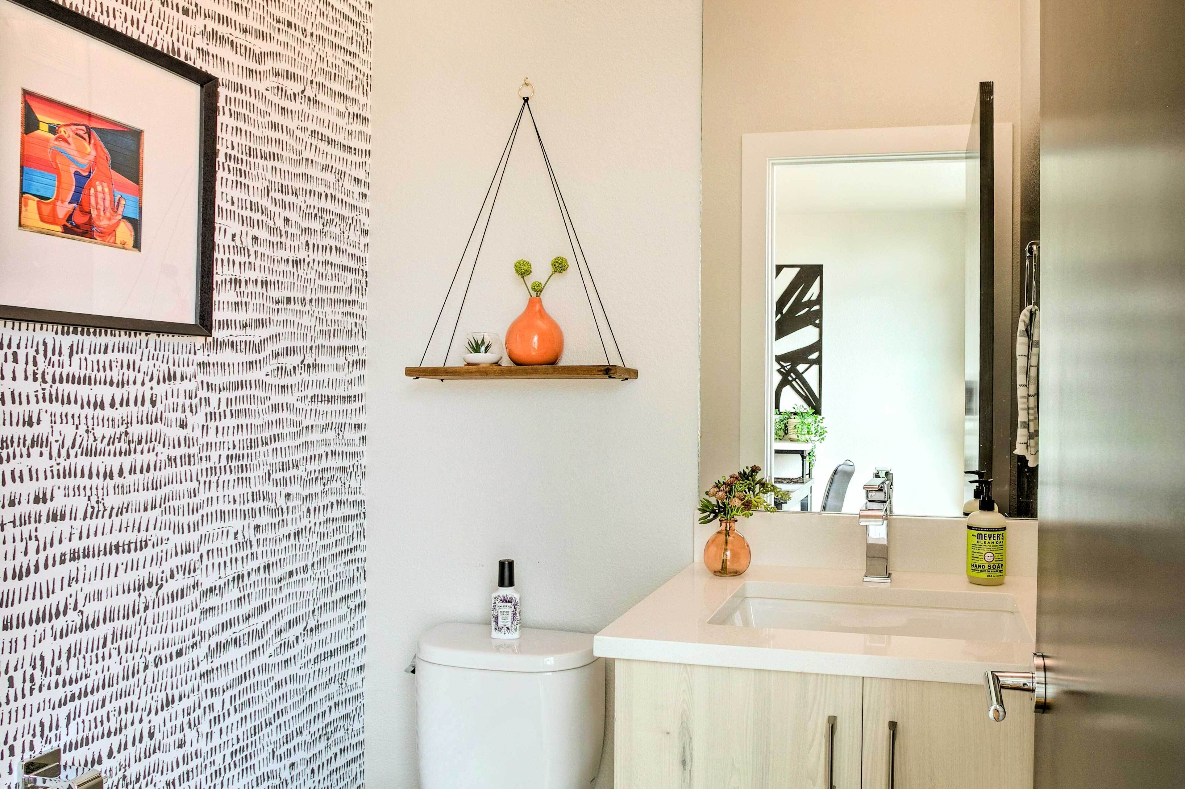 A bathroom with a patterned wall, a framed artwork, a hanging shelf with a vase, a toilet, and a sink with a mirror.