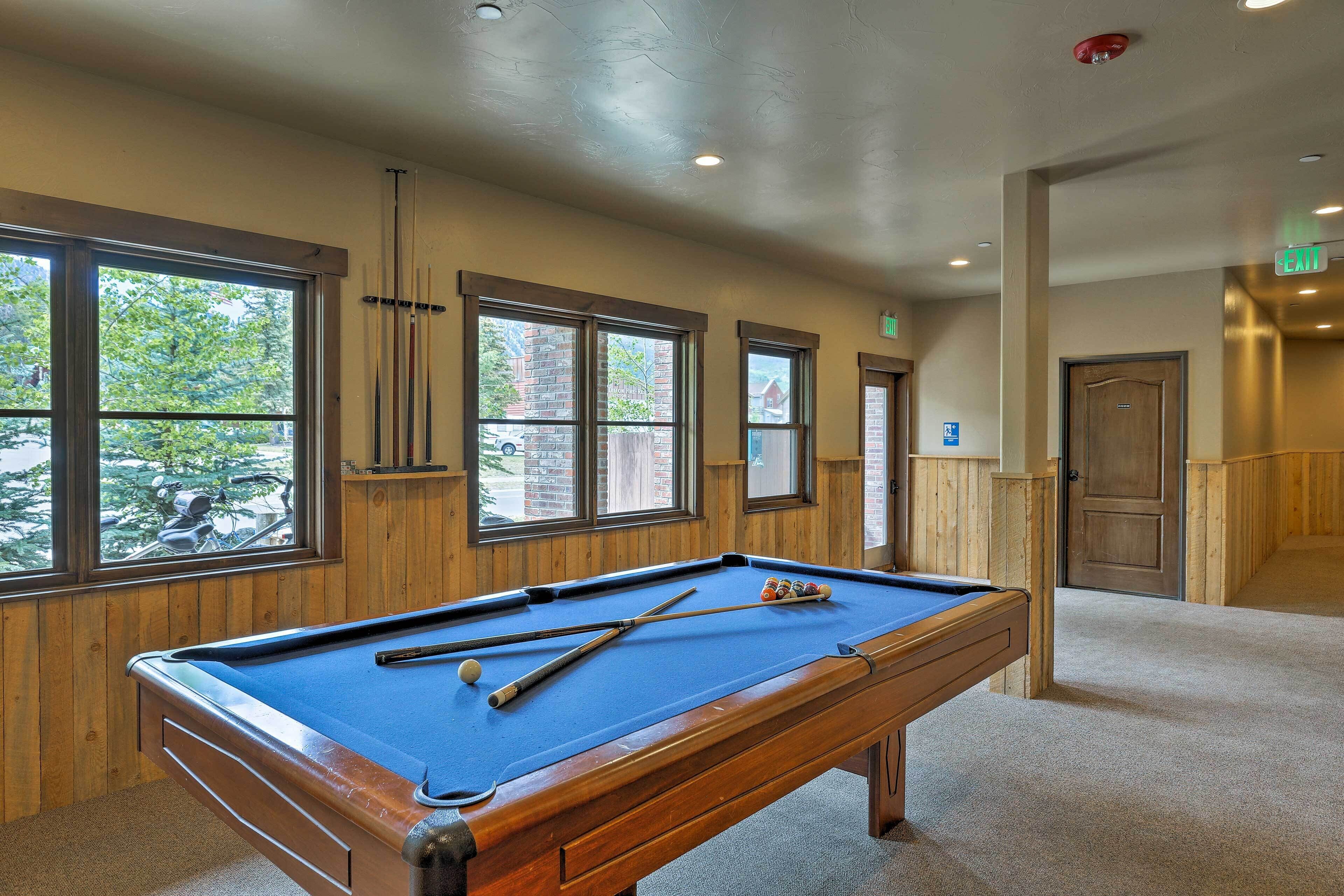 A pool table in a room with large windows, wooden walls, and a carpeted floor.