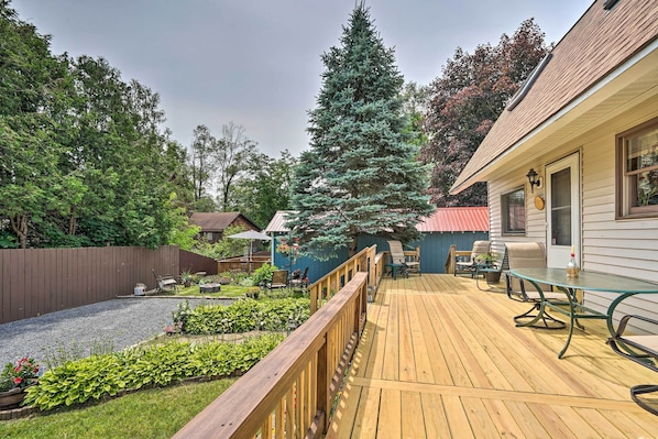 A wooden deck with outdoor furniture, a tall evergreen tree, and a house with a red roof. - Bolton Landing Romantic Getaway for Two (Bolton Landing)