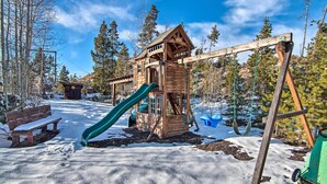 A wooden play structure with a slide and swings, surrounded by snow and trees. - Outdoor Kitchen & Swim Spa: 1-acre Grand Lake Home (Grand Lake)