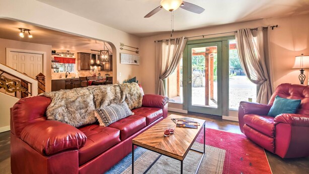 A spacious living room with a red leather sofa, a wooden coffee table, and a red armchair. A ceiling fan and a kitchen area are visible in the background.