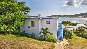 A small white building with a blue door and windows, surrounded by greenery and overlooking a body of water with boats. - Tropical Oasis: Bay Views ~ 2 Mi to Playa Sardinas (Culebra)