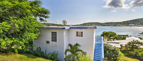 A small white building with a blue door and windows, surrounded by greenery and overlooking a body of water with boats.