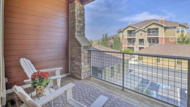 A balcony with a white Adirondack chair and a flower vase, overlooking a parking area and a multi-story building with stone accents. - Ski, Hike & Golf: Granby Condo w/ Shared Amenities (Granby)