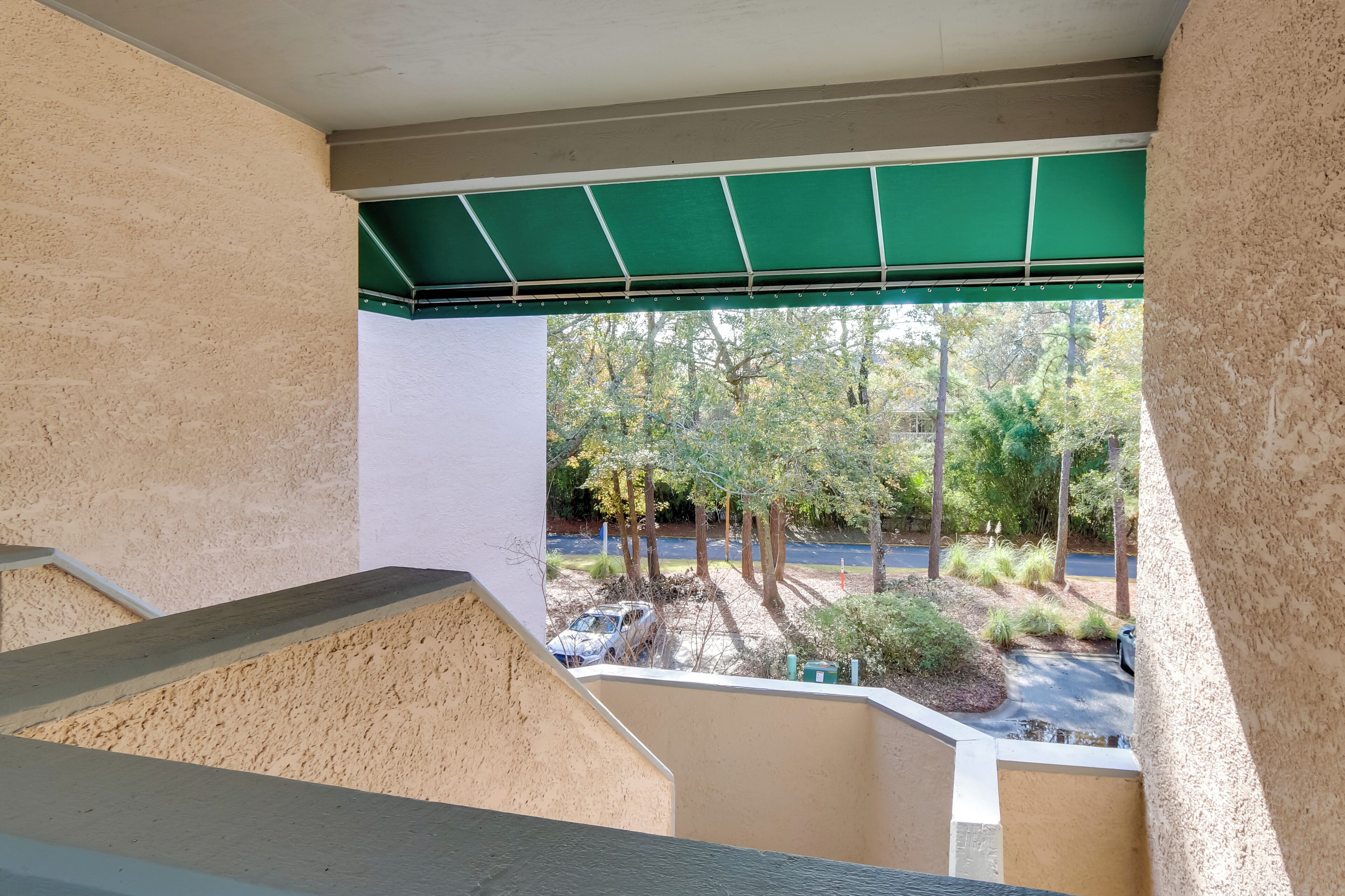 A view from a balcony with a green awning, a parking area with cars, and trees in the background.