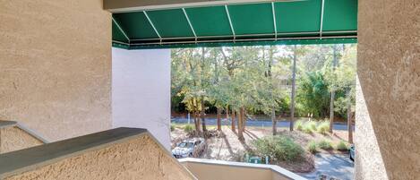 A view from a balcony with a green awning, a parking area with cars, and trees in the background.
