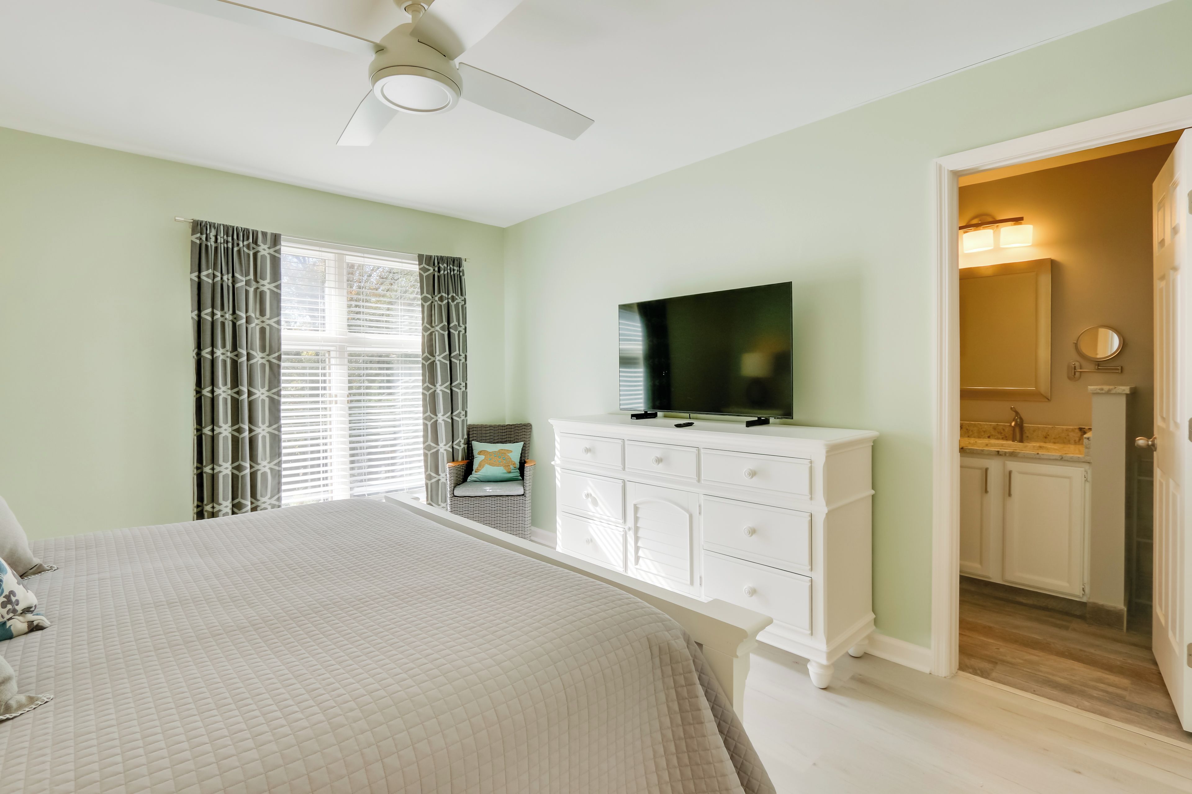 Un dormitorio con cama, televisor, cómoda, silla y baño a la vista a través de una puerta abierta.