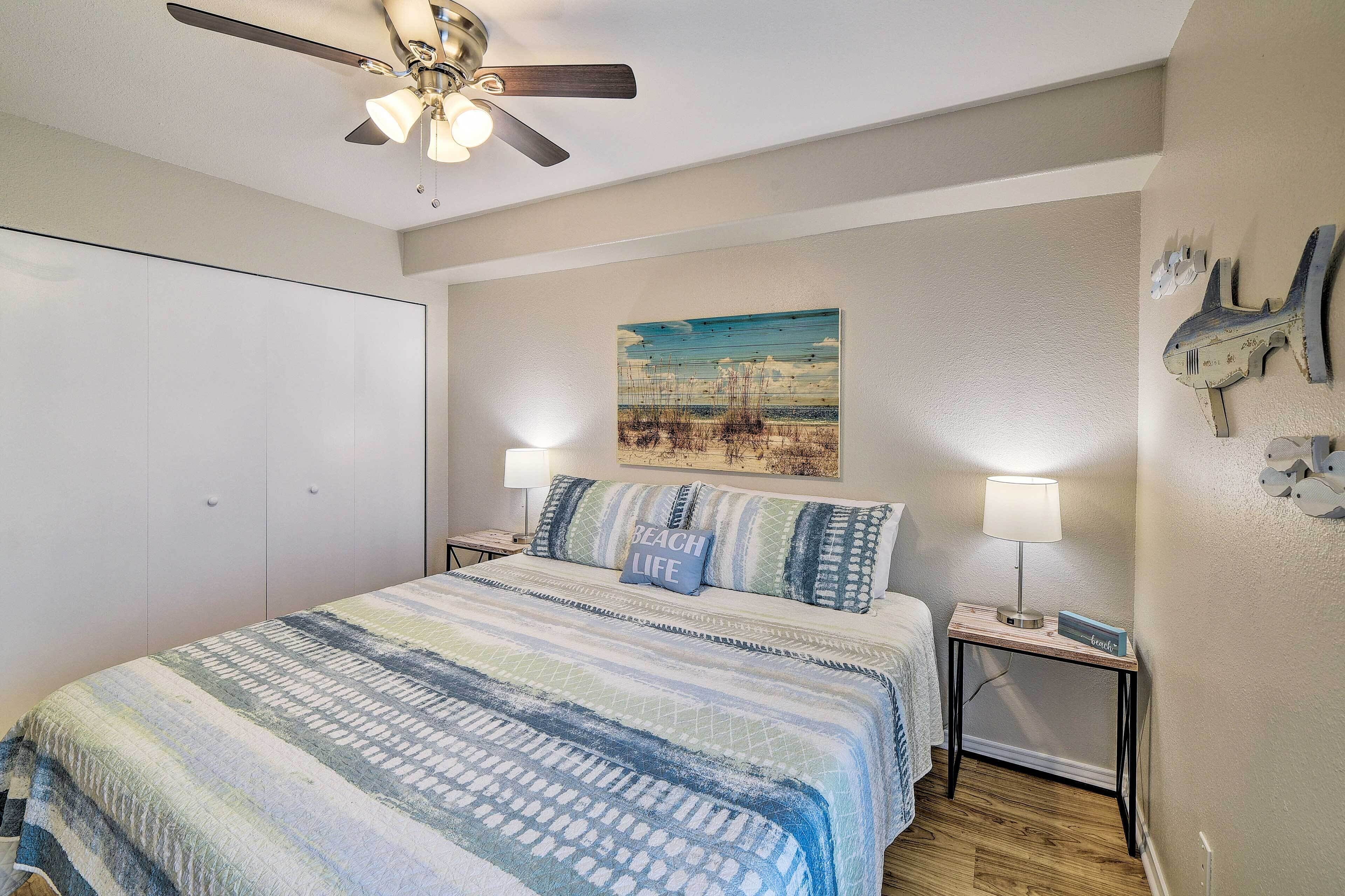 A bedroom with a bed, a ceiling fan, a nightstand with a lamp, and a wall painting.
