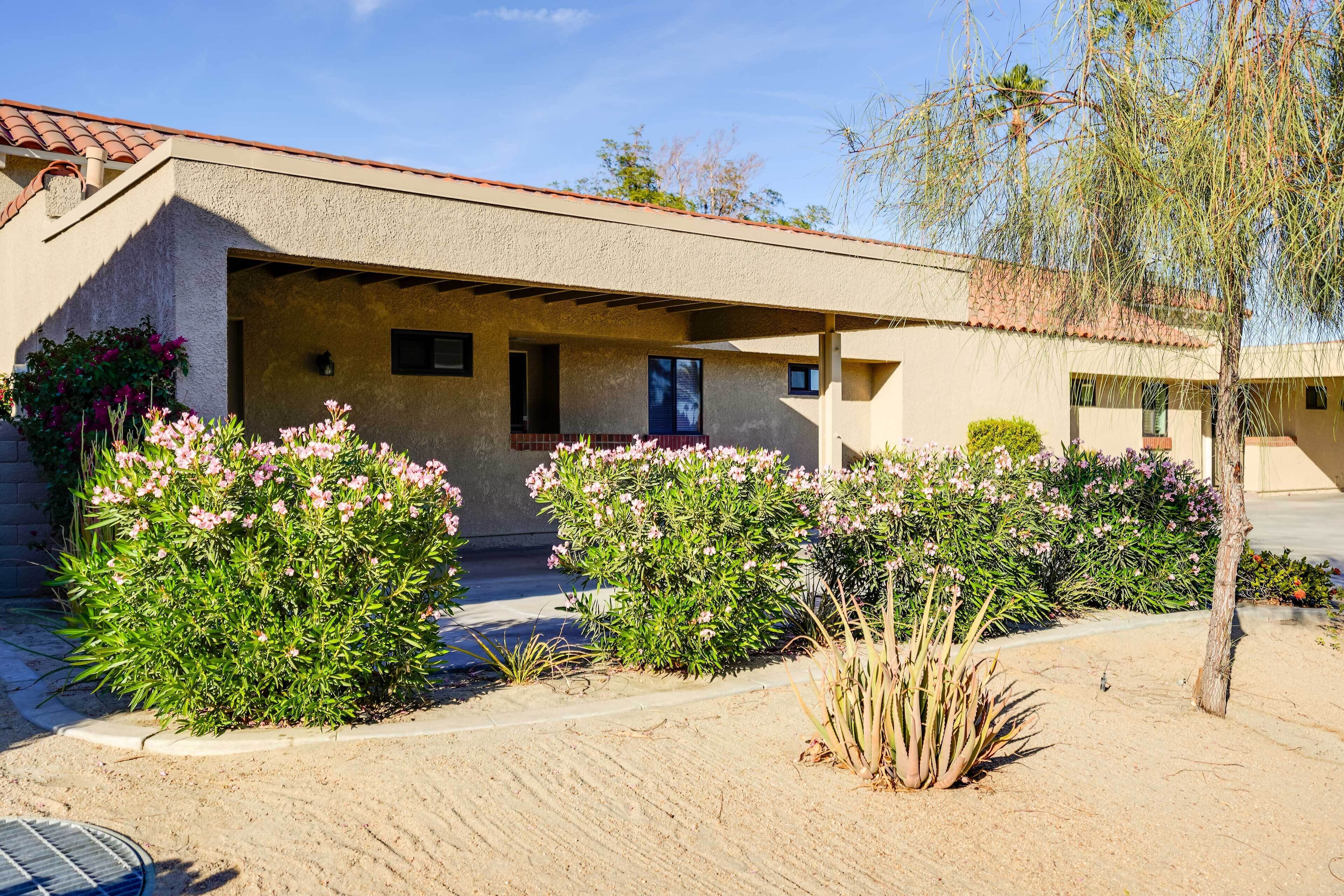A stucco building with a tiled roof, a covered porch, and a landscaped garden with pink flowering plants.