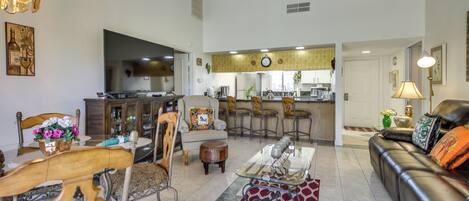 A spacious living room with a sofa, a coffee table, and a dining area with chairs and a table. There is a kitchen in the background with a refrigerator and a microwave.