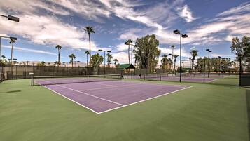 Una cancha de tenis al aire libre con superficie verde, rodeada por una valla, y palmeras al fondo.