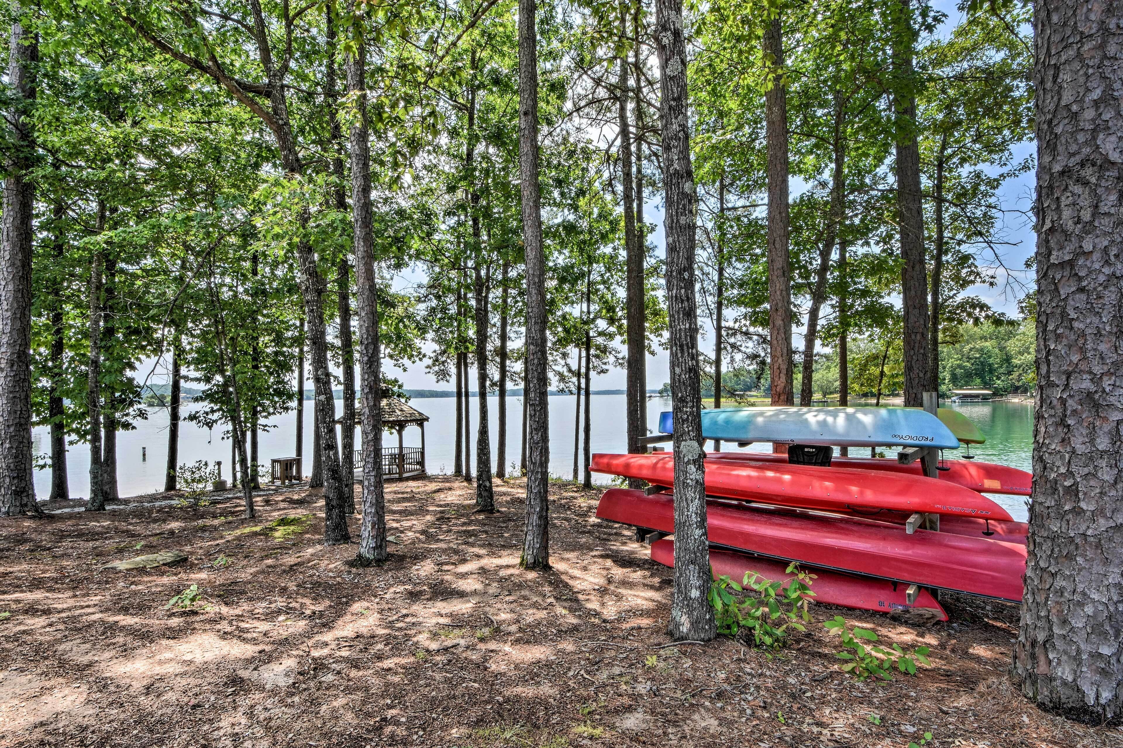 A lakeside setting with a row of red canoes and a blue canoe tied to a tree, surrounded by tall trees and a dock in the background.