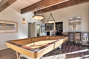 Une table de billard dans une pièce aux poutres apparentes, un coin bar avec des verres, et une œuvre d’art encadrée accrochée au mur.