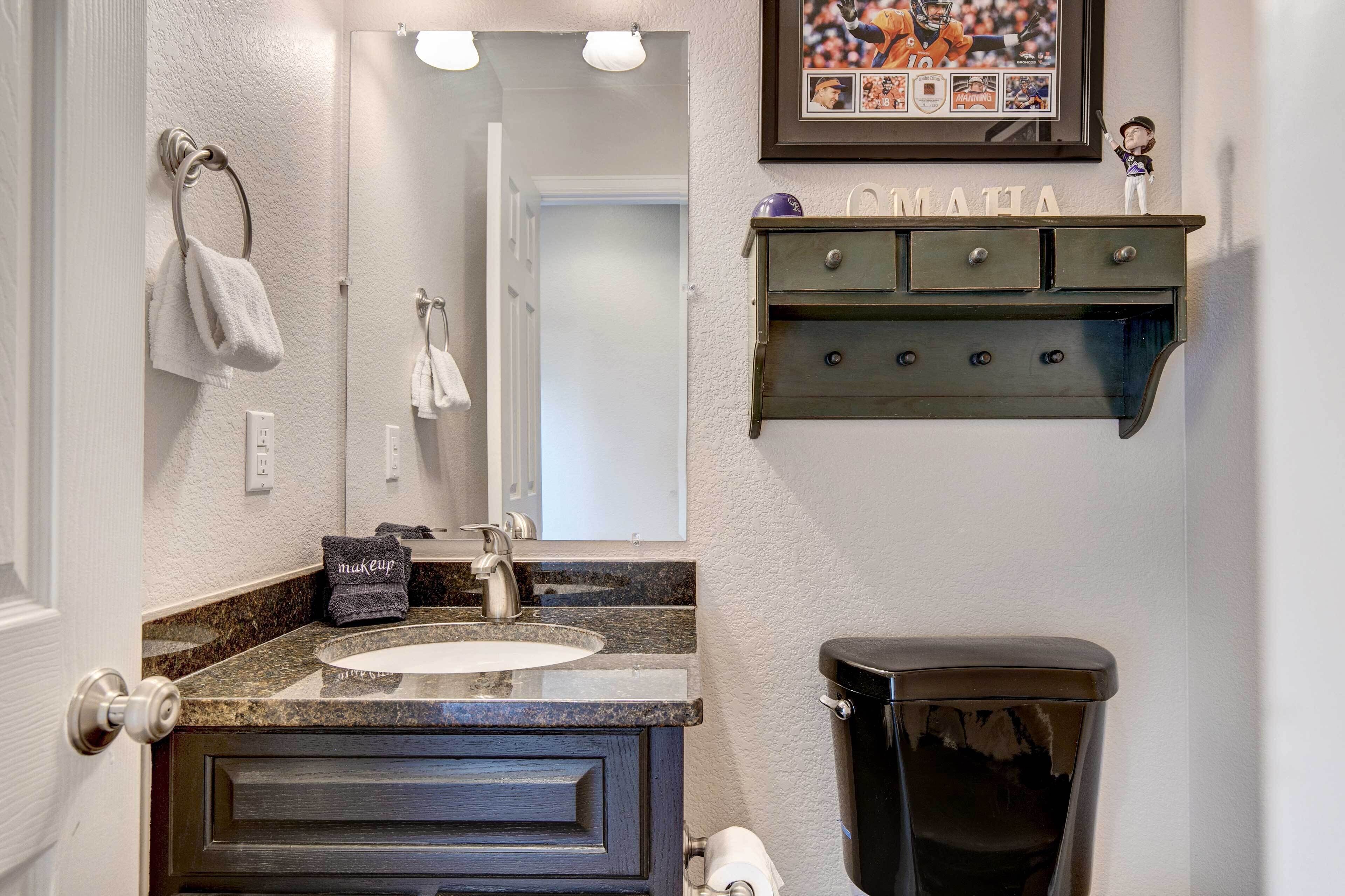 Une salle de bain avec un lavabo sur un comptoir en granit, un meuble-lavabo en bois foncé, des toilettes noires, un porte-serviettes avec une serviette blanche, un cadre contenant des objets de collection sur le thème du sport et une petite étagère sur laquelle est inscrit le mot « MATHA ».