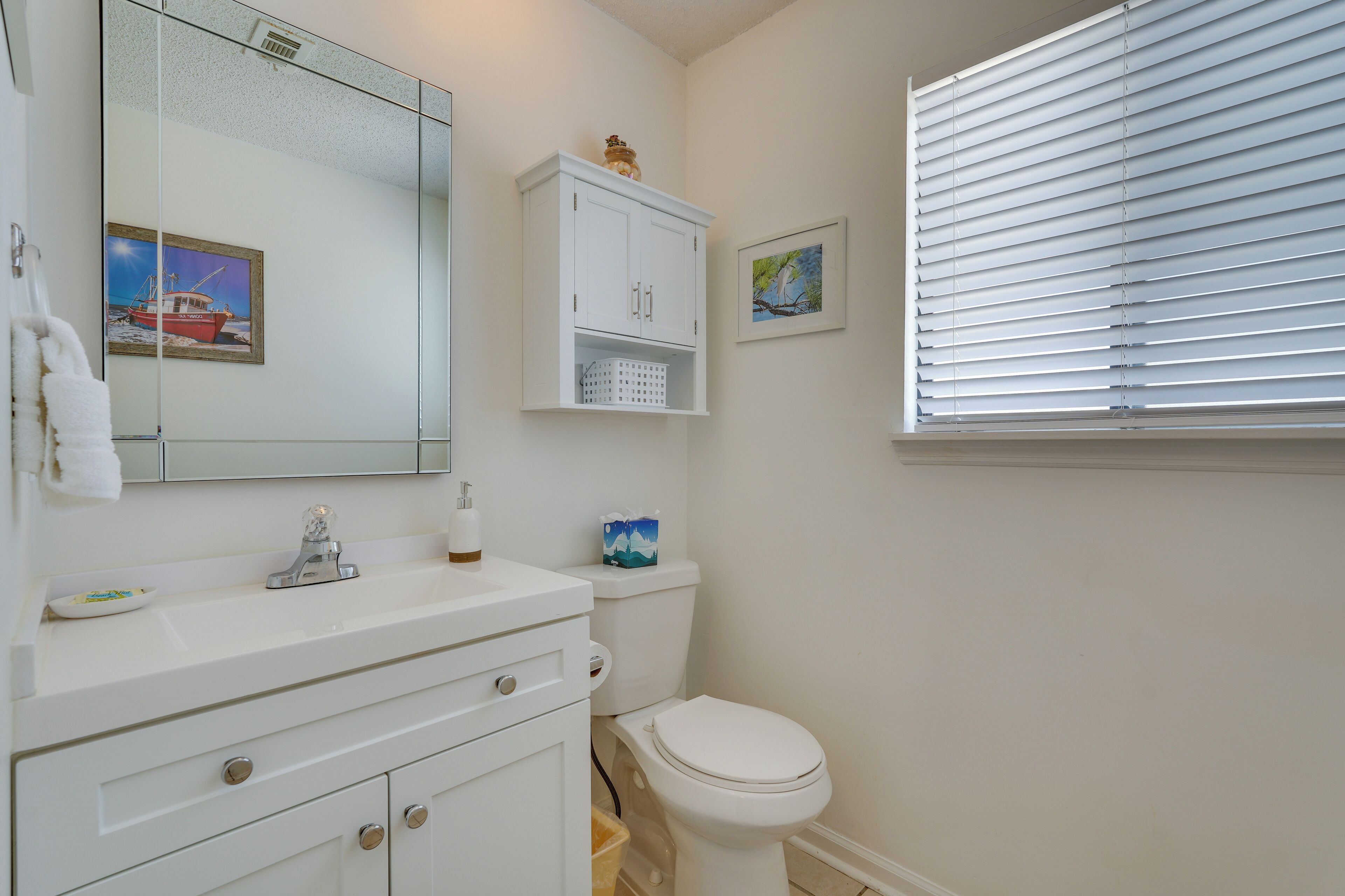 A bathroom with a white sink, mirror, and toilet. There are two framed pictures on the wall, a white cabinet above the toilet, and a window with blinds.