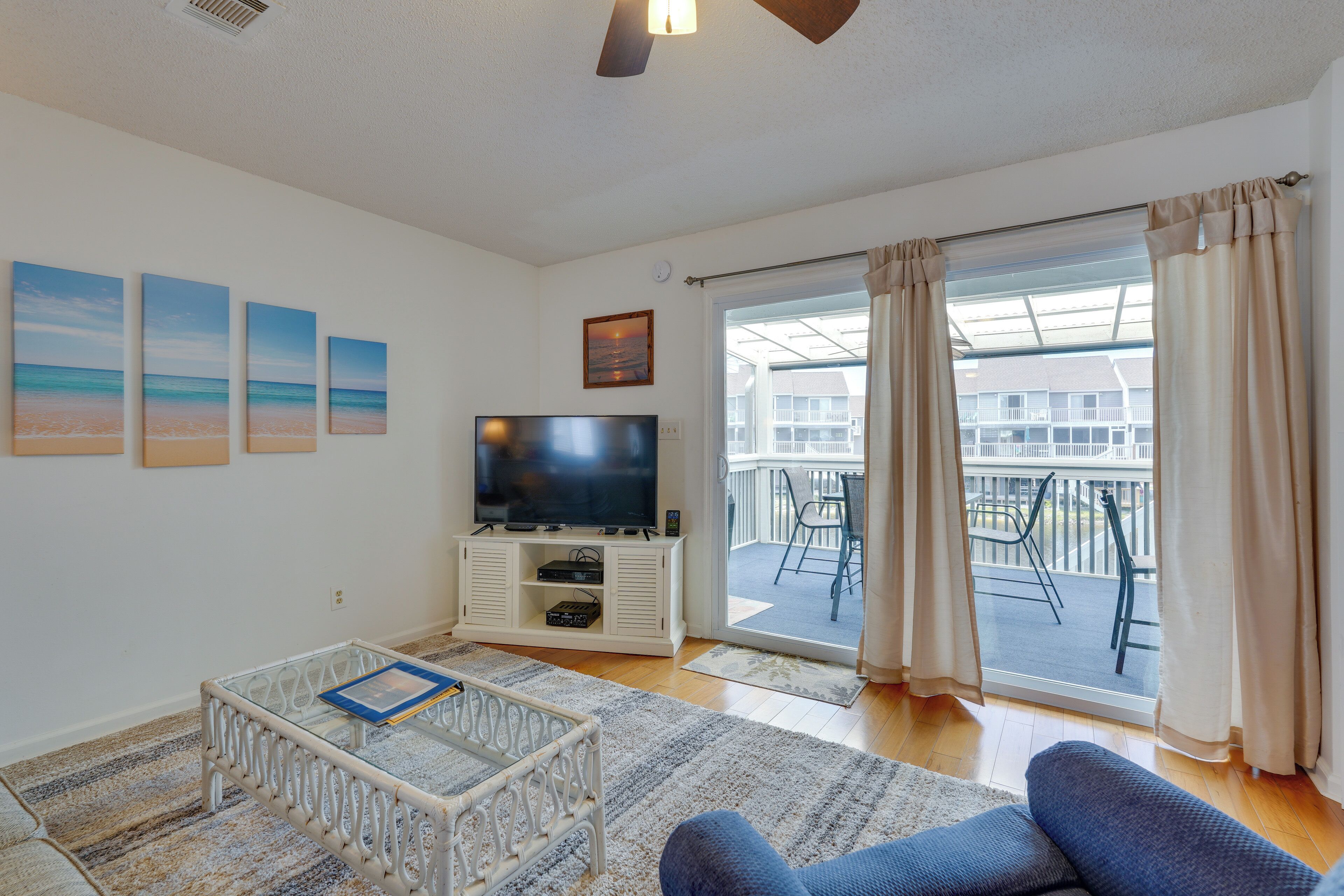 A living room with a flat-screen TV, a coffee table, and a sofa. There are beach-themed paintings on the wall and a balcony with a view.