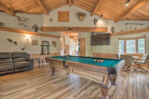 A pool table in a room with wooden floors, a leather sofa, and a television mounted on the wall.