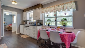 A kitchen with white cabinets, a dining table with a pink tablecloth, and a window with potted plants. - Norman Home w/ Yard, Walk to Park & OU Campus! (Norman)