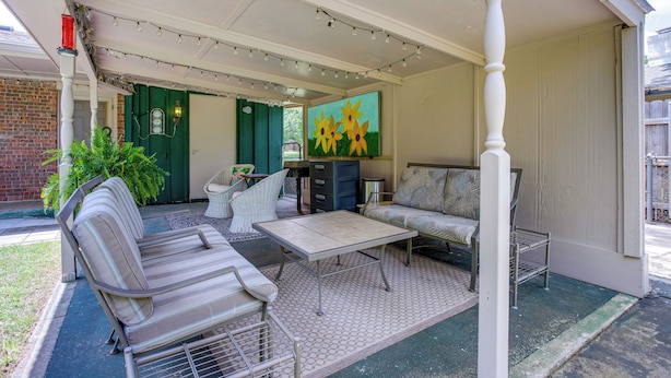 A covered patio area with a sofa, a table, and a chair. There is a painting of sunflowers on the wall, and a green door is visible in the background. - Norman Home w/ Yard, Walk to Park & OU Campus! (Norman)