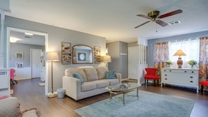 A living room with a beige sofa, a glass coffee table, a red armchair, a white dresser, and a ceiling fan. - Norman Home w/ Yard, Walk to Park & OU Campus! (Norman)