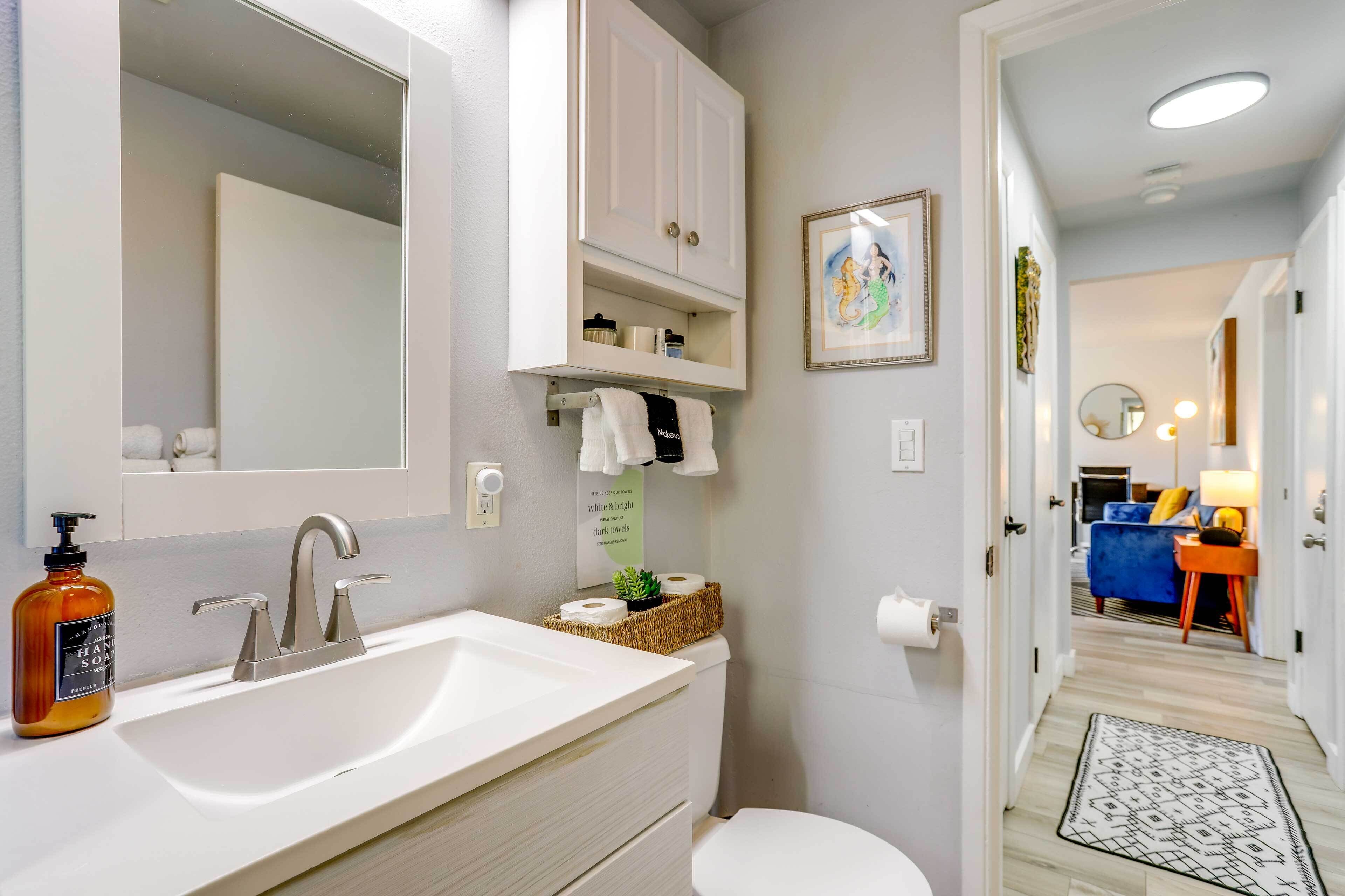 Une salle de bain moderne avec un lavabo blanc, un miroir et des armoires. On y trouve un porte-rouleau de papier toilette et une petite plante sur le comptoir. Un couloir mène à une chambre avec un lit bleu et une table de chevet.