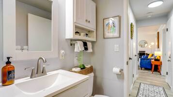 Une salle de bain moderne avec un lavabo blanc, un miroir et des armoires. On y trouve un porte-rouleau de papier toilette et une petite plante sur le comptoir. Un couloir mène à une chambre avec un lit bleu et une table de chevet.