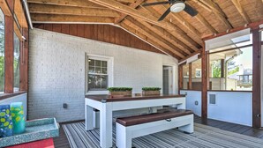 Un patio techado con techo de madera, pared de ladrillo blanco, una mesa con banco y plantas en macetas.