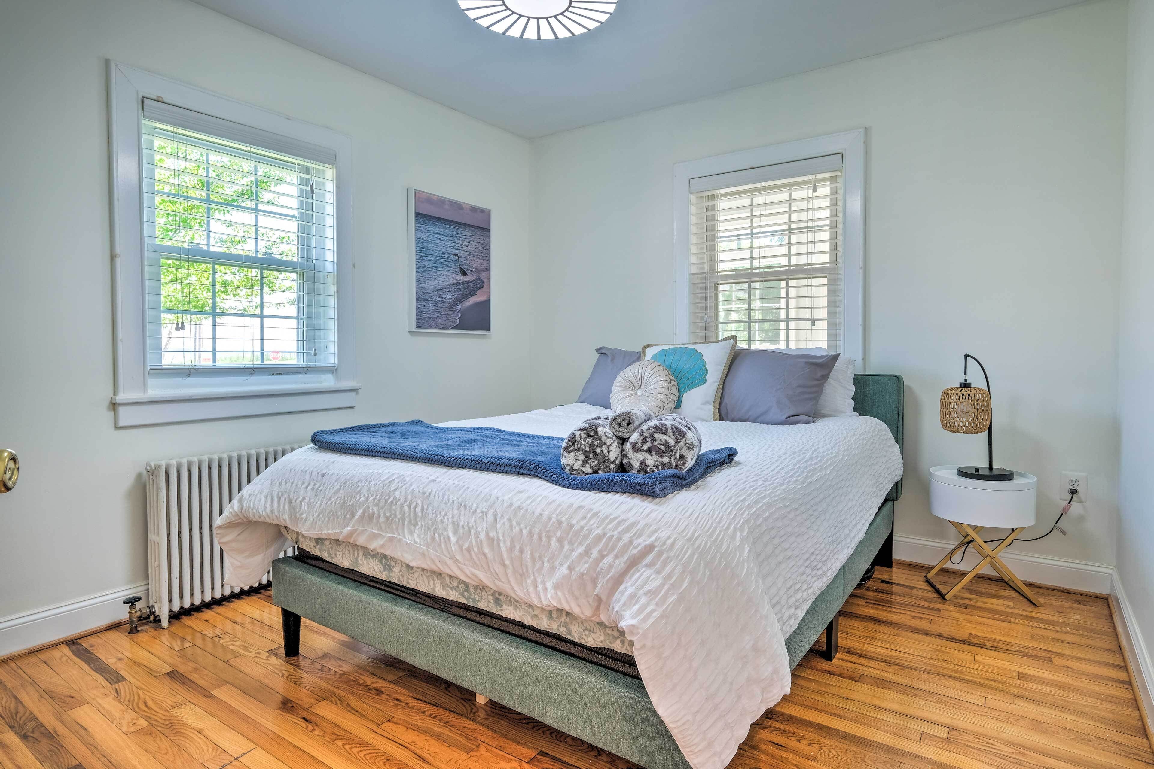 A bedroom with a bed, two windows, a radiator, a bedside table with a lamp, and a wall-mounted picture.