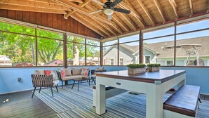 Un patio exterior con una mesa, sillas, un ventilador de techo y ventanas amplias con vistas a una zona residencial.
