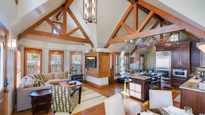 A spacious living area with a high ceiling, wooden beams, a fireplace, a television, and a kitchen area with modern appliances. - Downtown Breckenridge Cottage: Walk to Main St! (Breckenridge)