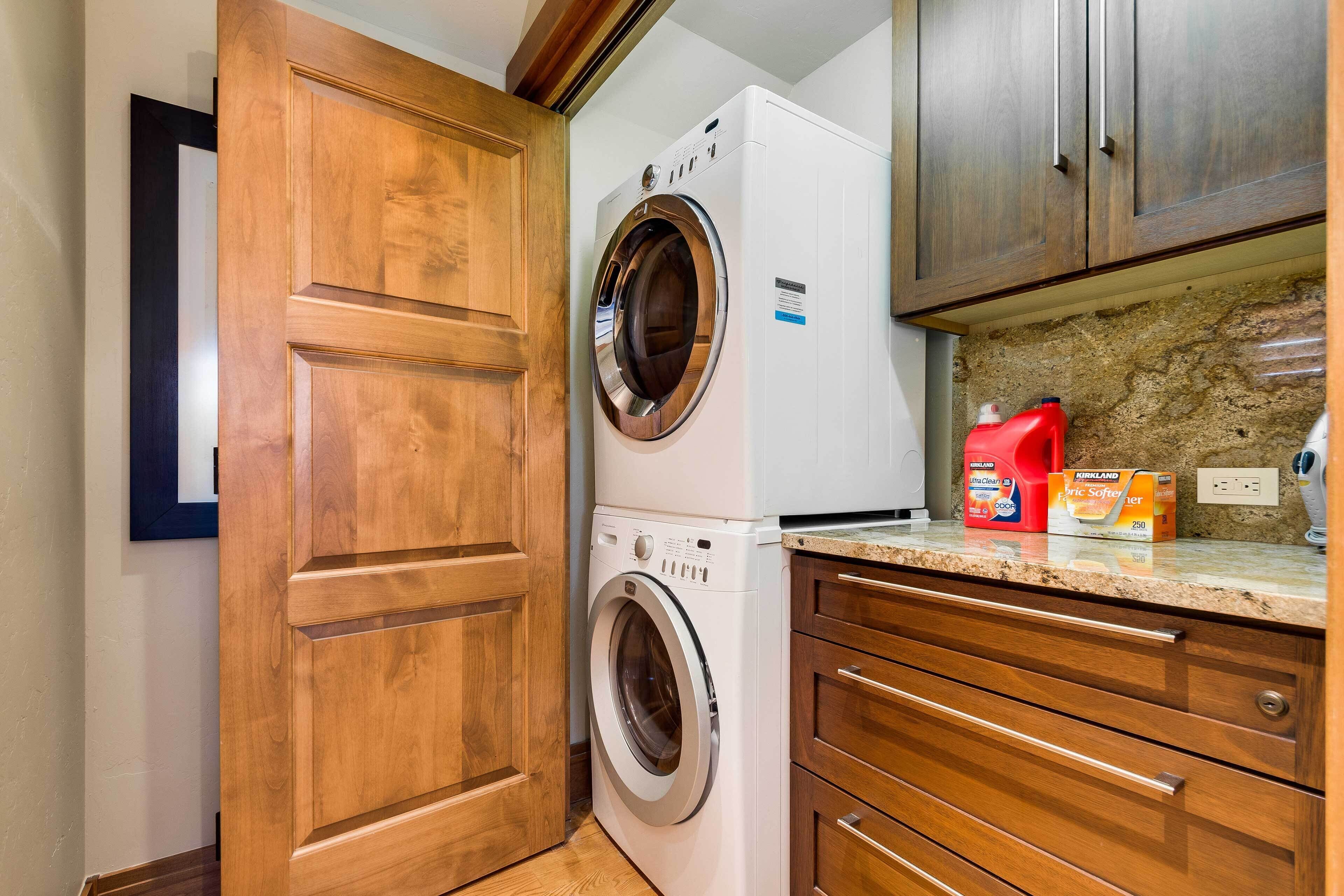 A laundry area with a front-loading washer and dryer, wooden cabinets, a granite countertop, and laundry detergent on the counter.