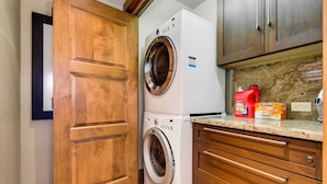 A compact laundry room with a front-loading washer and dryer, wooden cabinetry, and a granite countertop with detergent bottles. - Downtown Breckenridge Cottage: Walk to Main St! (Breckenridge)