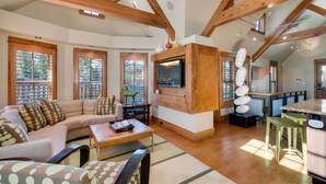 A spacious living area with a wooden ceiling, a large TV mounted on a wooden wall, a sofa with patterned cushions, a coffee table, and a dining area with stools. - Downtown Breckenridge Cottage: Walk to Main St! (Breckenridge)