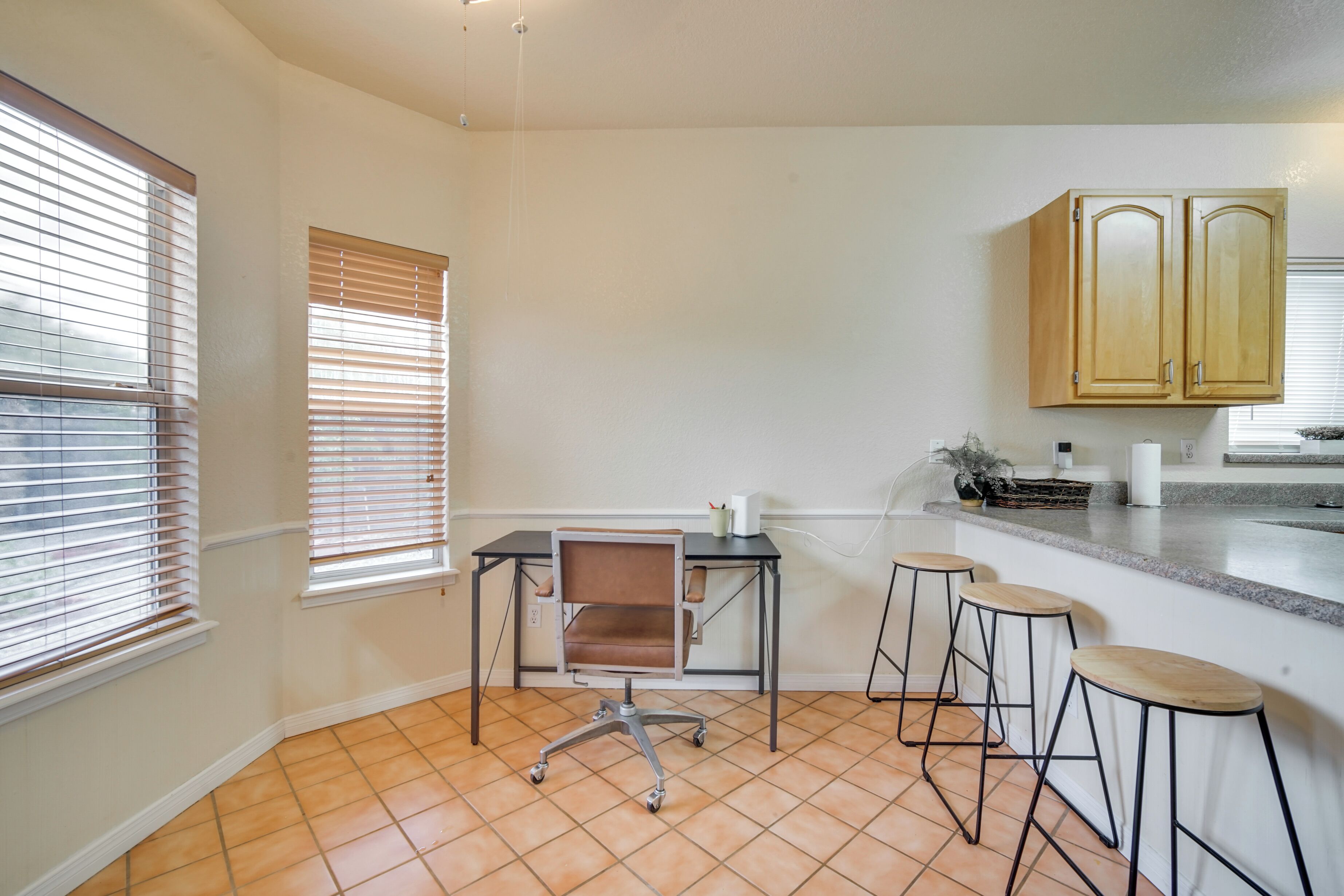 A kitchen with a dining area, a window with blinds, a chair, and bar stools.