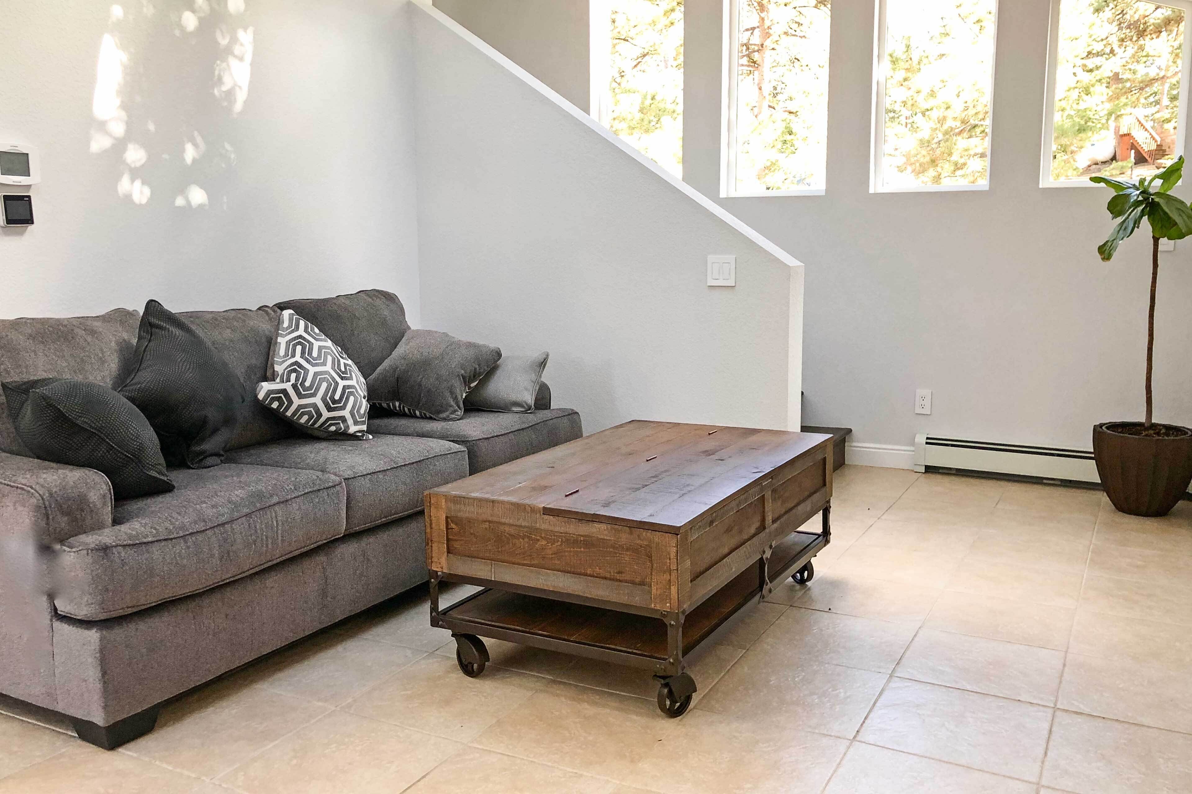 Una sala de estar con un sofá gris, una mesa de centro de madera con ruedas y un ventanal con vistas a árboles.