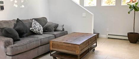 Una sala de estar con un sofá gris, una mesa de centro de madera con ruedas y un ventanal con vistas a árboles.