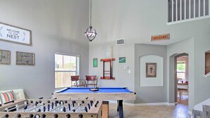 A room with a foosball table, pool table, and a billiards table. There are signs on the walls, a window, and a door leading to another room.