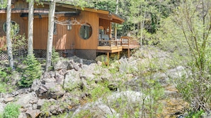 Une cabane en bois dotée d’une terrasse, entourée d’arbres et bordée d’un ruisseau aux rives rocheuses.