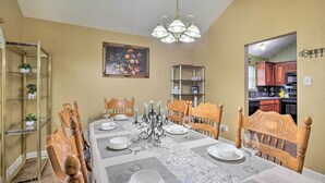 A dining room with a table set for four, surrounded by wooden chairs. There is a chandelier above the table, a painting on the wall, and a kitchen visible through an open doorway.