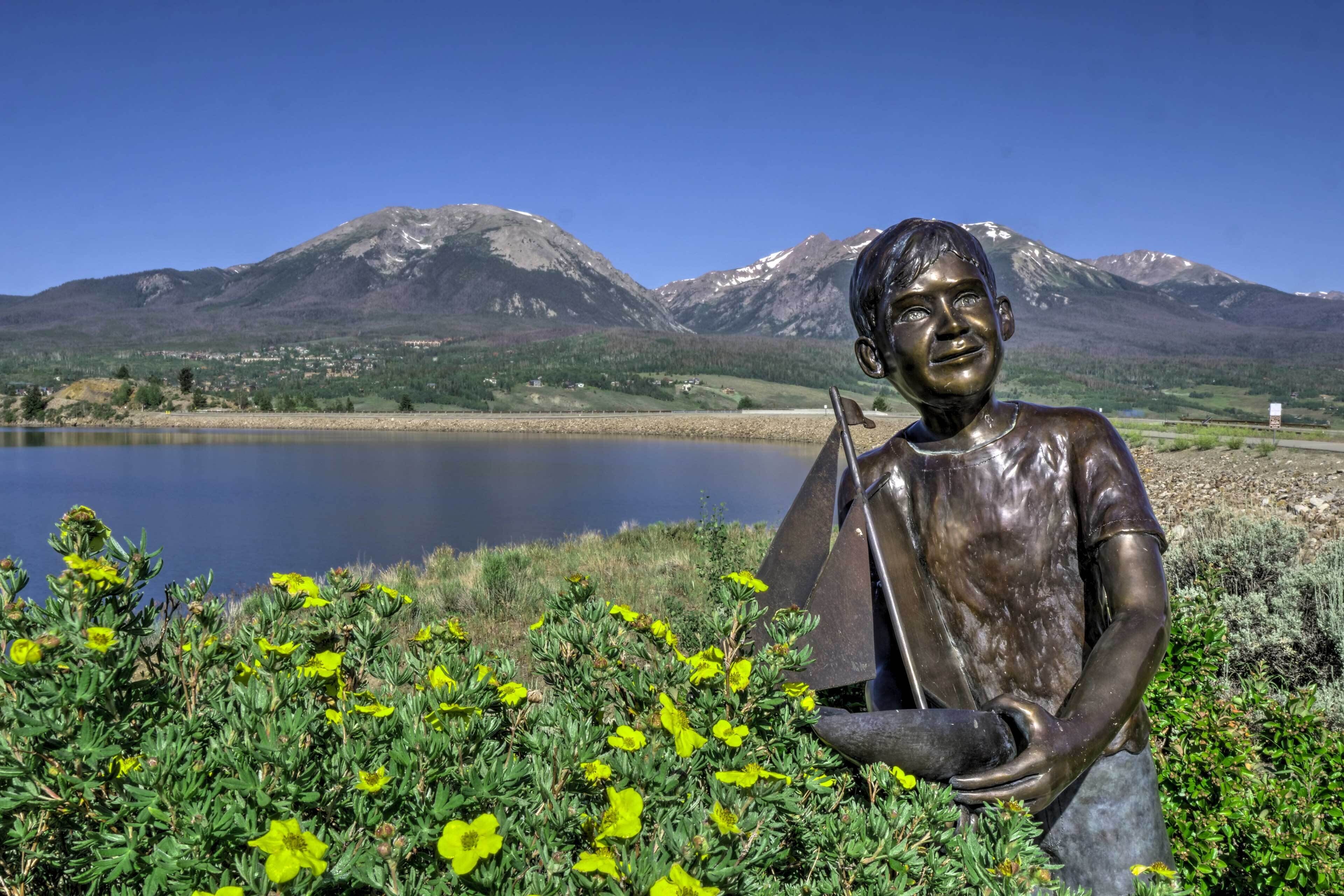 Una estatua de una persona con una mochila en un entorno natural, con montañas y un lago al fondo.