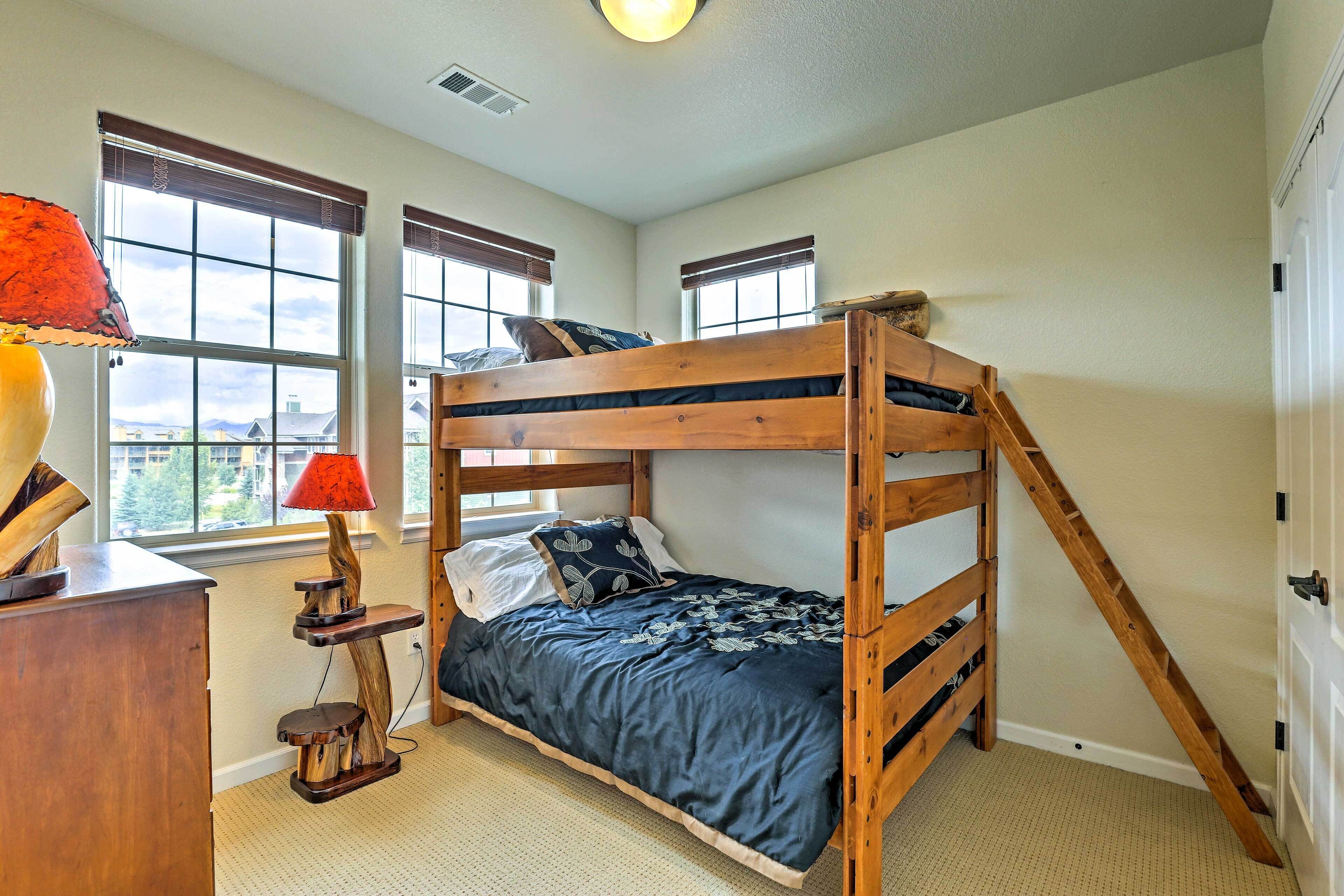 A bunk bed room with a desk and lamp, large windows, and a view of buildings outside.