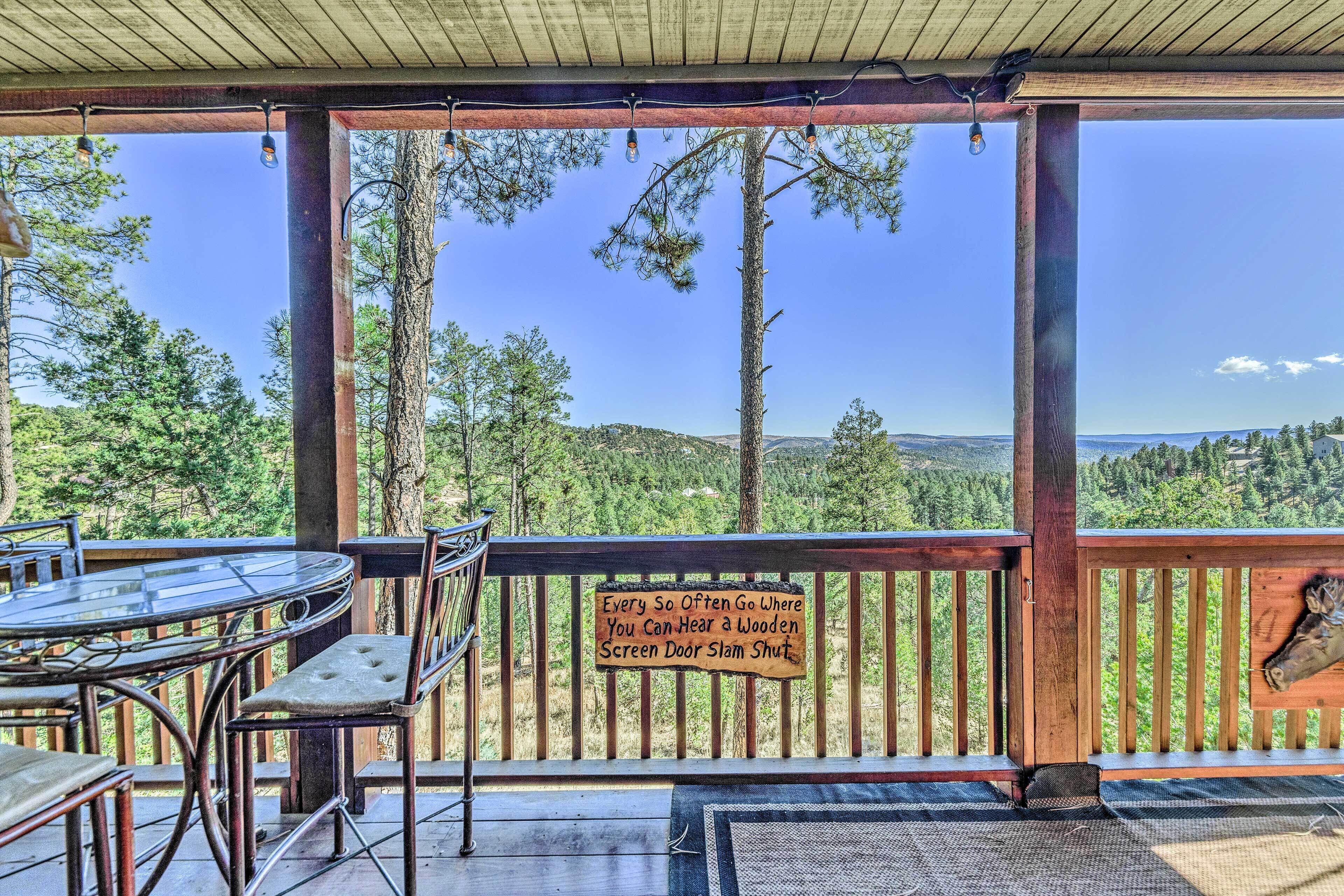 Una terraza de madera con una mesa y sillas de vidrio, con vistas a un paisaje boscoso.