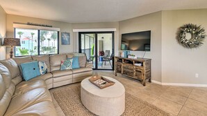A living room with a beige sectional sofa, a round ottoman, a flat-screen TV, and a wooden entertainment center.