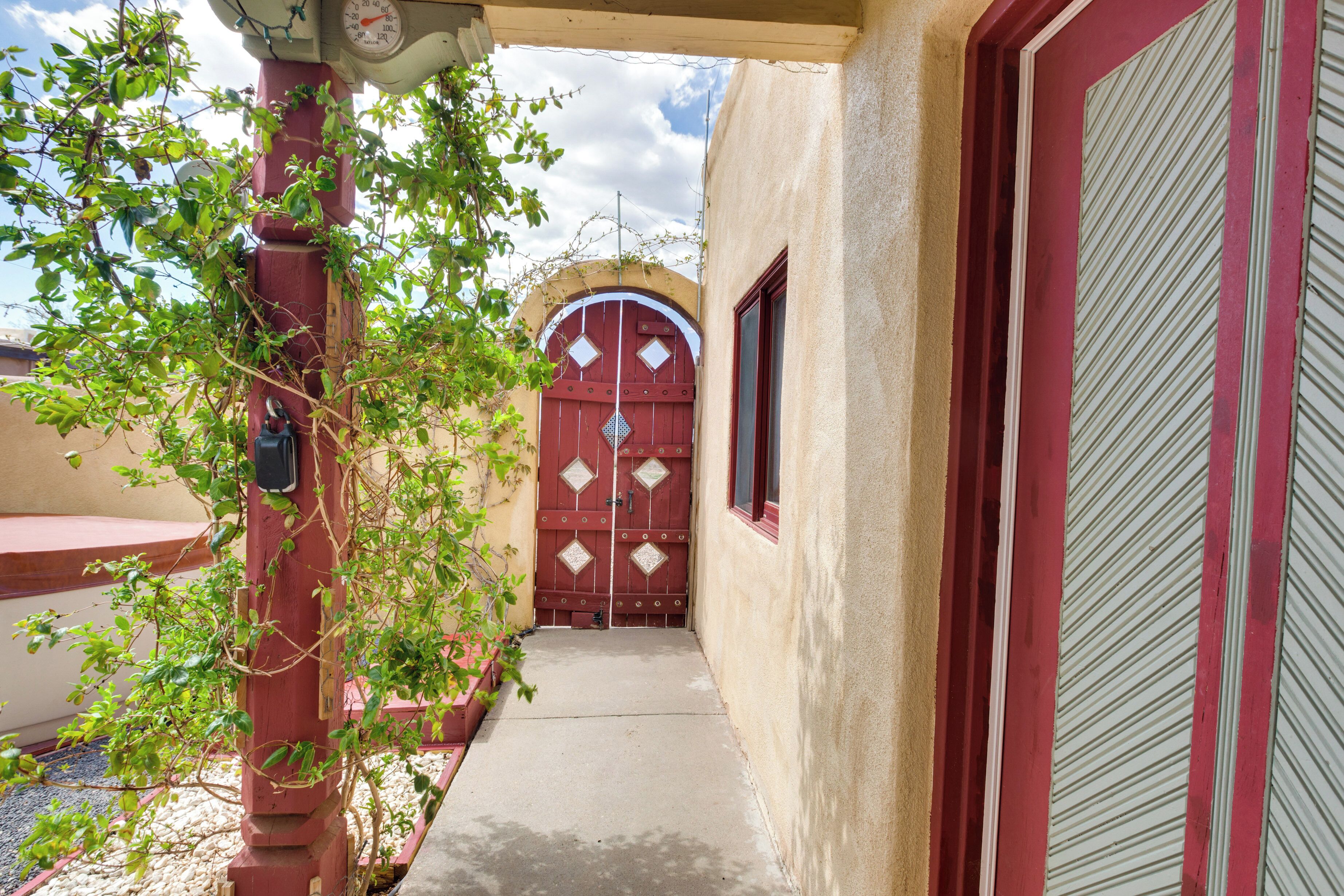 A narrow pathway leading to a red door with diamond patterns, flanked by a red pillar and a window with a striped blind.