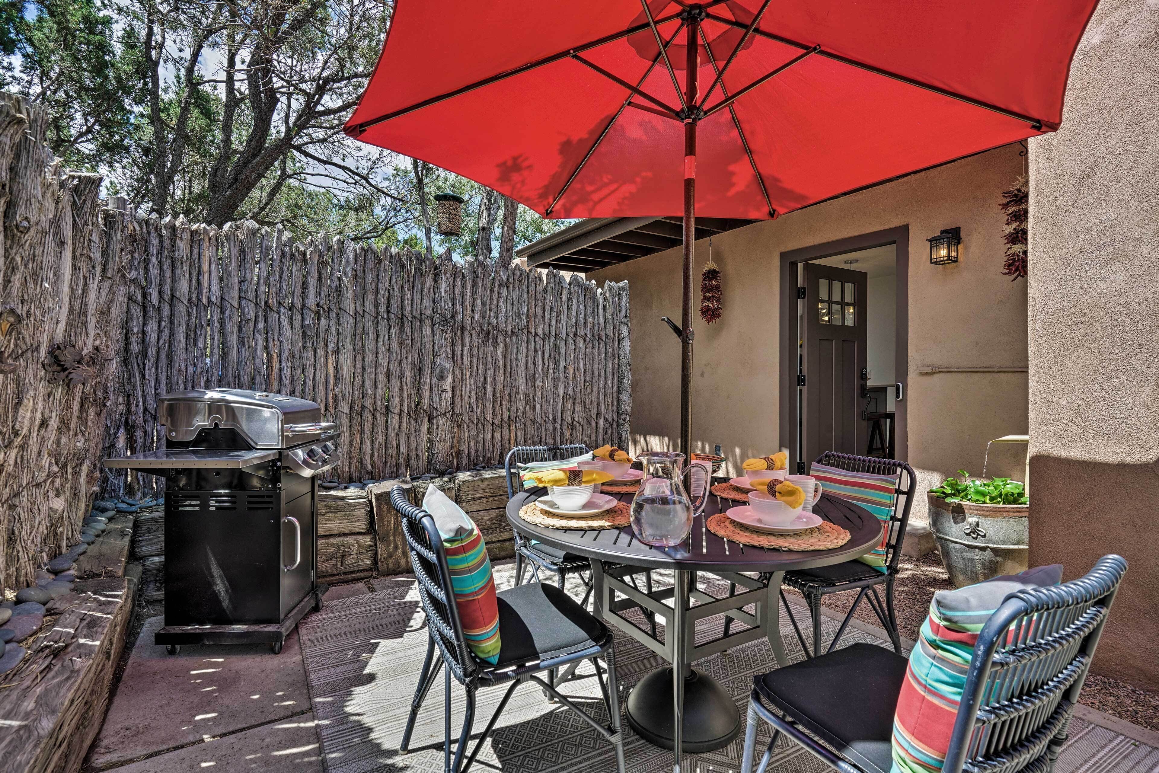Une terrasse extérieure avec une table ronde, des chaises, un barbecue et un parasol rouge.
