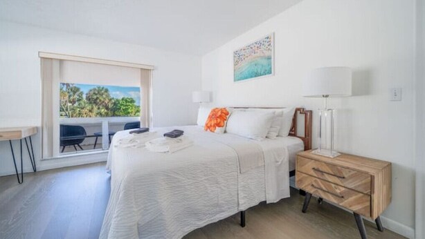 Un dormitorio con cama, escritorio, silla, mesita de noche y ventana con vista a árboles.