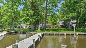 Une jetée en bois s’étendant sur une étendue d’eau calme, entourée d’arbres verdoyants et de deux maisons dotées d’une pelouse bien entretenue.