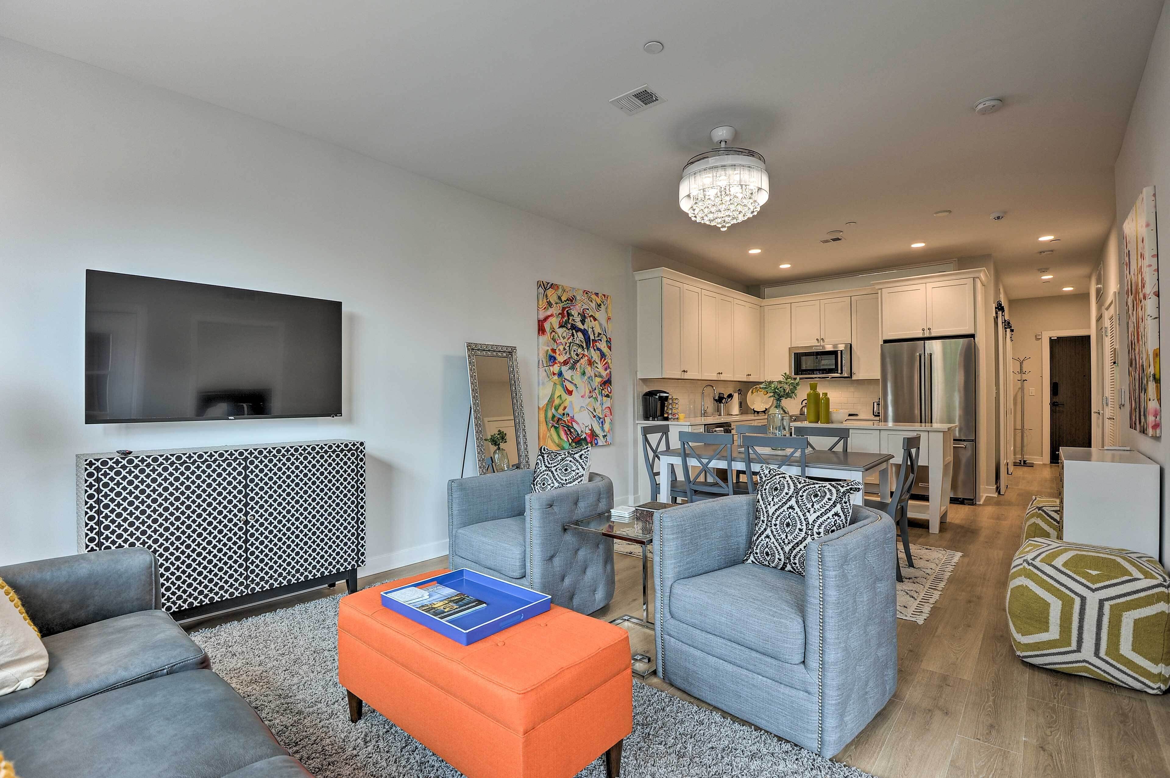 A modern living room with a flat-screen TV, grey sofas, an orange ottoman, and a patterned radiator.