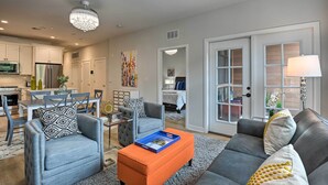 A modern living room with a grey sofa, a blue armchair, and an orange ottoman. There is a dining area with a table and chairs, and a kitchen with white cabinets and a refrigerator. - Chic Condo w/ Balcony in the Heart of Annapolis! (Annapolis)